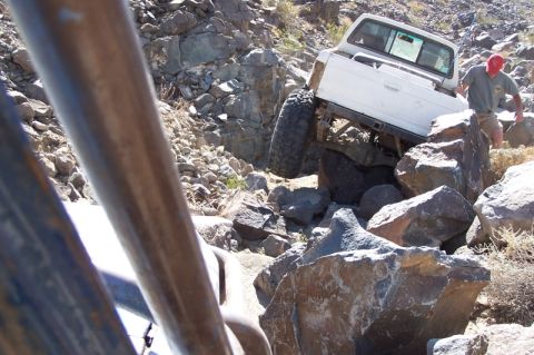 1990 Ford Bronco - A white pickup truck is carefully maneuvering over large rocks on a , desert-like terrain.
