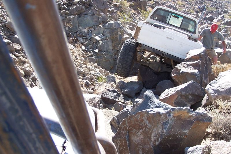 White pickup truck navigating a rocky, off-road trail with a person walking nearby