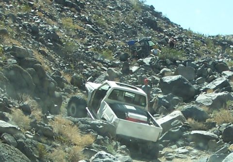1990 Ford Bronco - Two off-road vehicles traverse a steep, rocky incline under a clear sky.