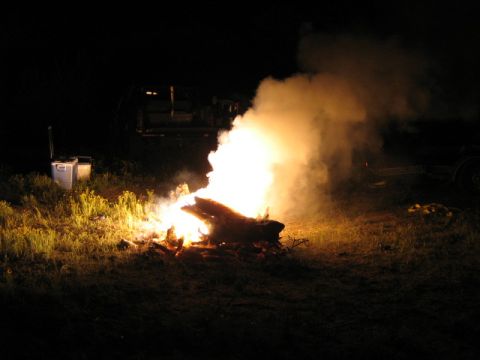 1986 Ford Bronco - A large bonfire burns brightly at night, casting an orange glow on the surrounding grass and smoke.