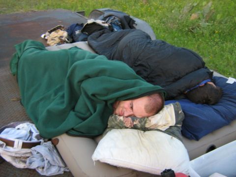 1986 Ford Bronco - Two individuals are seen sleeping outdoors, covered by blankets and sleeping bags on an air mattress.