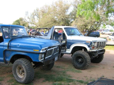 1986 Ford Bronco - Two modified Ford trucks, a blue pickup and a white/blue Bronco, parked outdoors on a dirt lot.