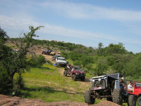 1986 Ford Bronco - A convoy of off-road vehicles navigates a challenging rocky trail surrounded by lush greenery and a bright blue sky.