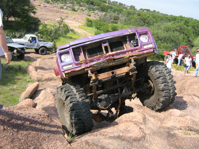 Purple off-road SUV with large tires navigating rocky terrain. Other trucks and people visible in background