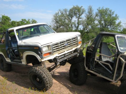 1986 Ford Bronco - Two modified off-road vehicles, a white Ford Bronco and a black custom build, are positioned closely together on a dirt path.
