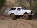 1994 Ford Bronco - This white Ford Bronco is navigating a dusty, off-road trail with large, mud-caked tires.