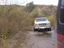 1994 Ford Bronco - A white Ford F-150 truck is seen driving on a wet dirt road with brush and a hill in the background.