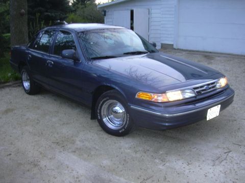 1996 Ford Crown Victoria - A dark blue sedan with chrome wheels is parked in front of a garage.