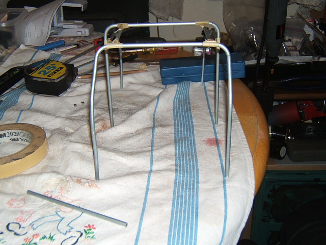 Metal frame with four legs, taped at the top corners, resting on a striped cloth. Tape measure and other items are nearby