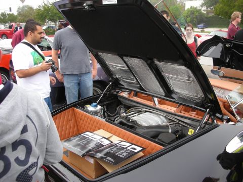 1998 Ford Crown Victoria - Spectators gather around a black supercar with its rear engine cover raised, revealing the engine and tan-trimmed bay.
