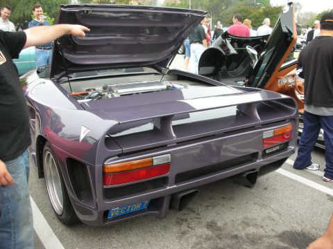 1998 Ford Crown Victoria - raised rear engine cover of a dark gray Vector supercar reveals its bay, with onlookers gathered around.