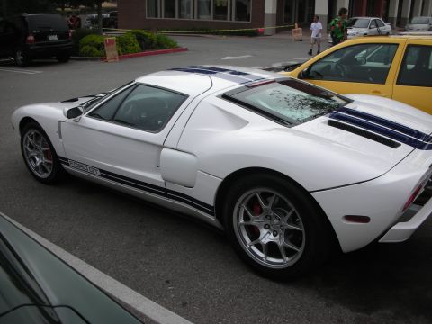 1998 Ford Crown Victoria - white Ford GT with twin blue racing stripes is parked in profile next to a yellow car at a show.
