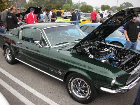 1998 Ford Crown Victoria - dark green classic Ford Mustang fastback sits hood-up showing its engine among rows of show cars and spectators.
