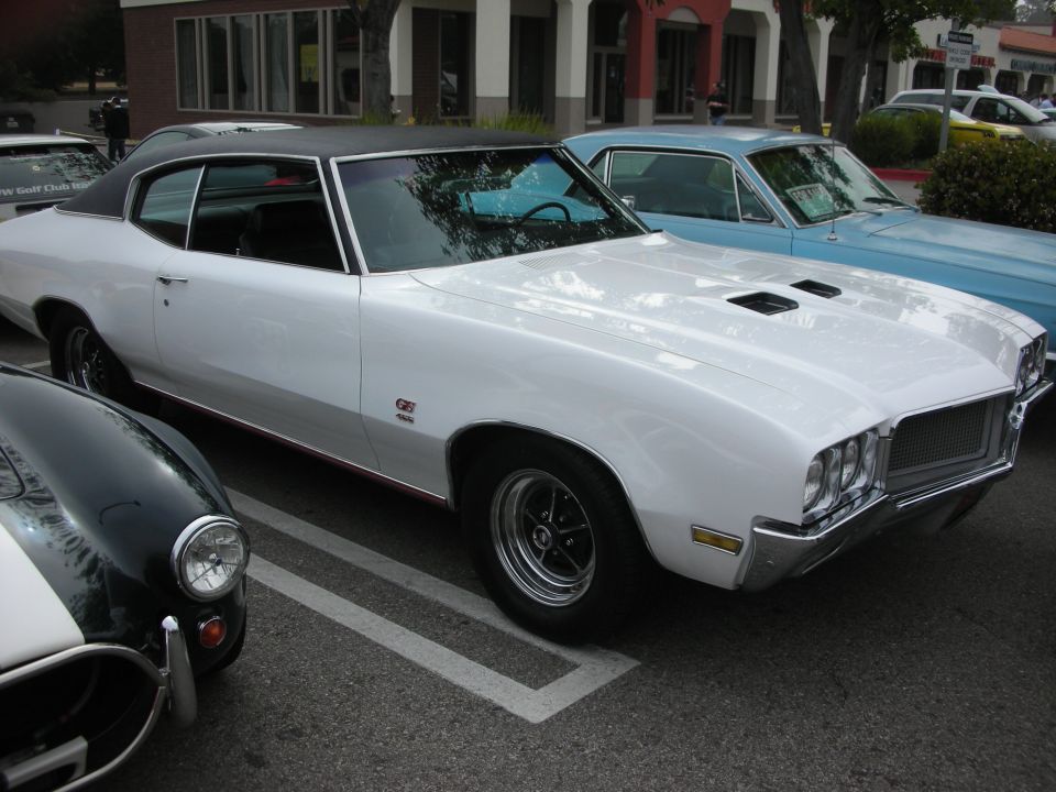 White Buick GS muscle coupe with hood scoops parked at a car show