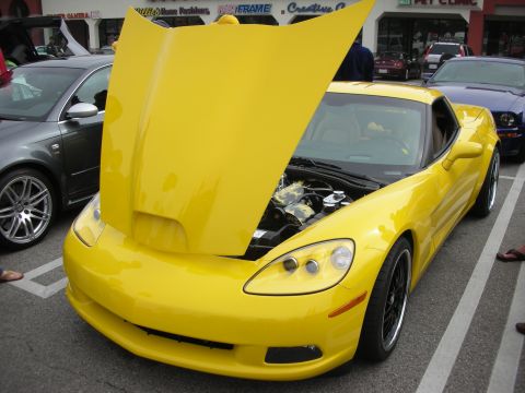 1998 Ford Crown Victoria - yellow Chevrolet Corvette C6 sits with its hood raised in a strip-mall parking lot car show.