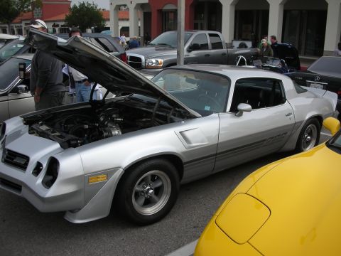 1998 Ford Crown Victoria - silver Camaro coupe parked with its hood raised at a busy outdoor car show, beside a yellow sports car.