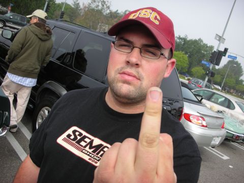 1998 Ford Crown Victoria - man wearing a maroon USC cap, glasses, and a black SEMA Show shirt raises his middle finger in a parking lot at a car event.
