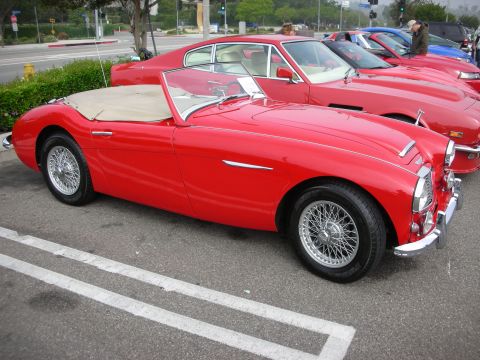 1998 Ford Crown Victoria - red vintage British convertible roadster with chrome wire wheels and a folded tan top, parked in a lot beside other classic cars.