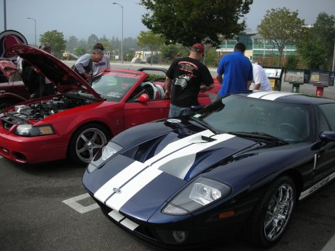 1998 Ford Crown Victoria - dark blue Ford GT with white racing stripes parked next to a red Mustang convertible with its hood up at a car meet, people milling around.