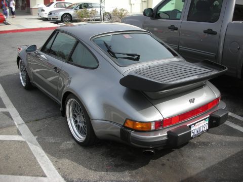 1998 Ford Crown Victoria - Rear three-quarter of a grey Porsche 911 Turbo with a whale-tail spoiler bearing a RUF badge, parked beside a pickup.