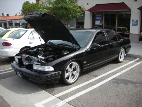 1998 Ford Crown Victoria - lowered black Chevrolet Impala SS sedan with its hood open and polished five-spoke wheels, parked in front of a storefront.