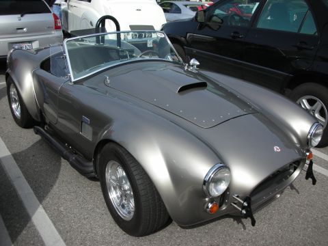 1998 Ford Crown Victoria - silver-grey Shelby Cobra-style roadster with chrome side pipes, hood scoop and polished wheels, parked among other cars.