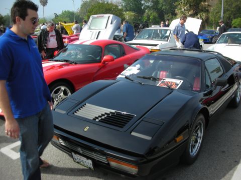 1998 Ford Crown Victoria - black Ferrari 328 with red interior and pop-up headlights at a car meet, a red Ferrari behind it and a spectator in a blue shirt.