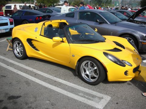 1998 Ford Crown Victoria - yellow Lotus Elise roadster with side intake scoops and polished wheels, shown front three-quarter in a crowded parking lot.