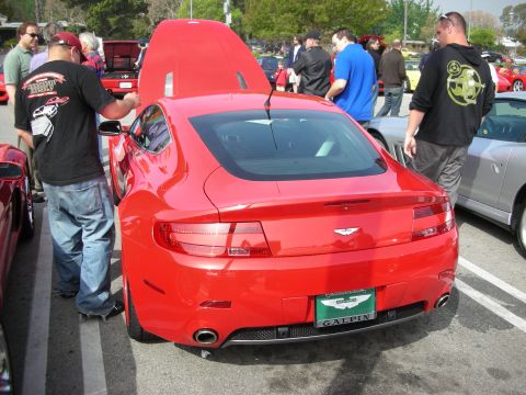 1998 Ford Crown Victoria - red Aston Martin V8 Vantage coupe shown rear-on with its rear hatch raised among parked cars and spectators at an outdoor show.