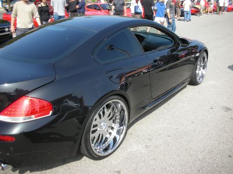 1998 Ford Crown Victoria - black BMW M6 coupe sits at an outdoor car meet, showing its rear quarter and oversized chrome multi-spoke wheels.