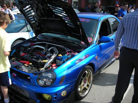 1998 Ford Crown Victoria - blue Subaru Impreza WRX with hood up reveals its boxer engine, wearing Falken livery and gold wheels at a car meet.
