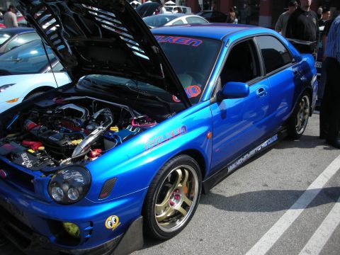 1998 Ford Crown Victoria - Side view of a blue Subaru Impreza WRX with its hood raised, gold wheels and Falken graphics down the body.