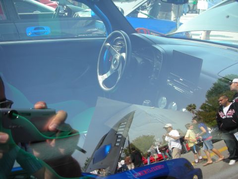 1998 Ford Crown Victoria - Looking through a windshield at a roadster cabin with a deep-dish steering wheel, reflections of the car show overlaid.