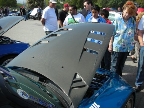 1998 Ford Crown Victoria - vented carbon-fiber hood stands open over a blue sports car as spectators gather around it at a car meet.