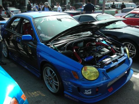 1998 Ford Crown Victoria - blue Subaru Impreza WRX with hood raised and yellow-tinted headlights, parked among cars at a busy meet.