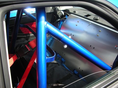 1998 Ford Crown Victoria - Close-up of a blue painted roll cage with diagonal bars, red harness and bare metal rear panel inside a race-prepped cabin.