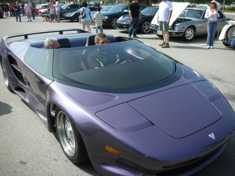 1998 Ford Crown Victoria - low purple convertible exotic sports car with two occupants seated inside, parked at a busy outdoor car meet.