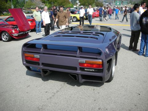 1998 Ford Crown Victoria - Rear view of a purple wedge-shaped exotic supercar with twin taillights and louvered engine cover, surrounded by onlookers.