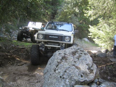 1987 Suzuki Samurai - Two off-road vehicles tackle a challenging, rocky path surrounded by trees.