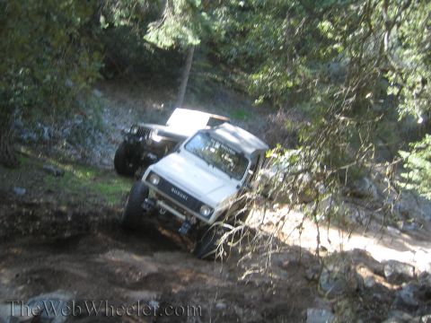 1987 Suzuki Samurai - A white 1987 Suzuki Samurai navigates a rocky, wooded trail, with another off-road vehicle following closely behind.