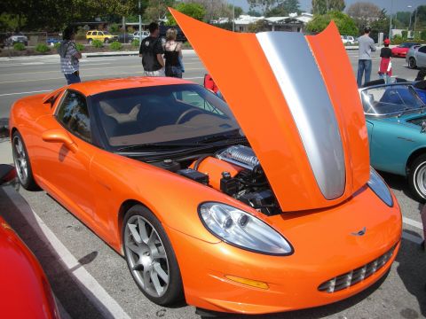 1998 Ford Crown Victoria - orange C6-era Corvette with its hood raised showing a silver center stripe, parked at a sunny outdoor car show.