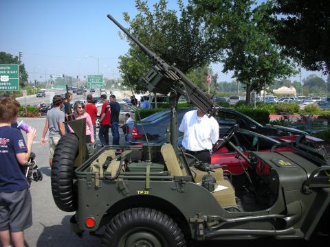 1998 Ford Crown Victoria - Olive-drab military Jeep with an upward-pointing mounted machine gun parked near a busy roadway with onlookers.