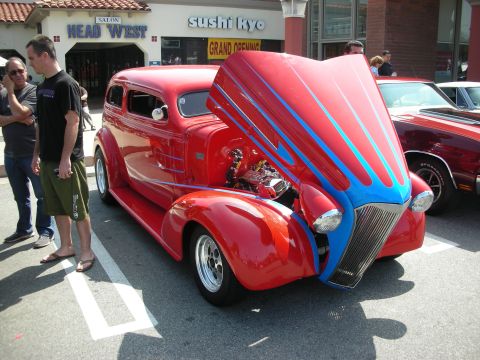 1998 Ford Crown Victoria - red 1930s-style street rod with blue flame graphics and its hood tilted up to show a chromed engine, parked outside a 'Sushi Kyo' storefron…