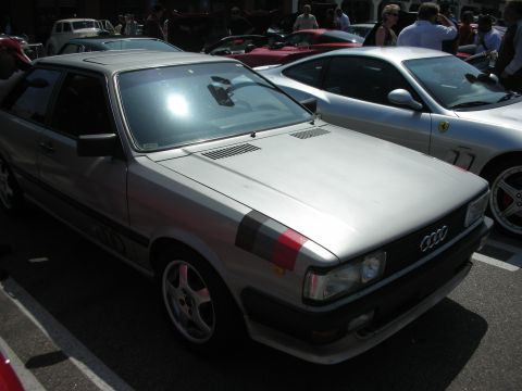 1998 Ford Crown Victoria - silver Audi coupe with a faded red-and-grey side stripe and the four-ring grille badge sits in a car-show lot beside a Ferrari.