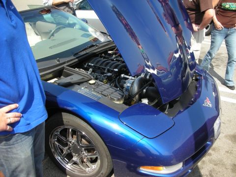 1998 Ford Crown Victoria - blue Corvette with its clamshell hood raised, revealing a V8 engine with a polished supercharger, beside chrome wheels at a meet.