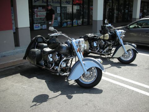 1998 Ford Crown Victoria - Two valanced-fender cruiser motorcycles, one black and one cream, parked at a curb in front of a photo store.