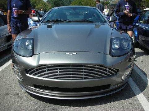 1998 Ford Crown Victoria - front end of a gray Aston Martin coupe with its winged badge and mesh grille, in a crowded car-show lot.