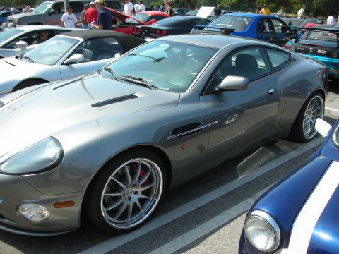 1998 Ford Crown Victoria - gray Aston Martin coupe shown in profile on multi-spoke wheels, parked among other cars at a show.