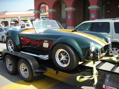 1998 Ford Crown Victoria - green Cobra-style roadster with a gold stripe strapped onto a flatbed trailer in front of a strip mall.