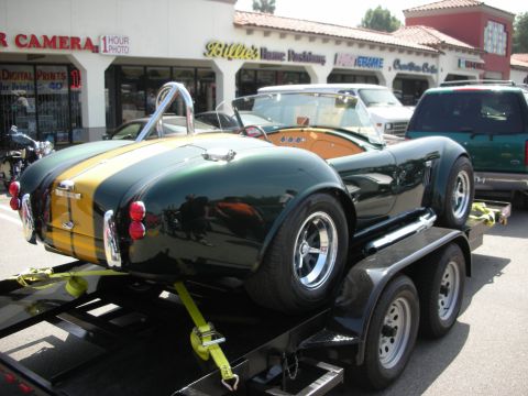 1998 Ford Crown Victoria - rear of a green Cobra-style roadster with a gold stripe strapped to a trailer near a camera and photo store.